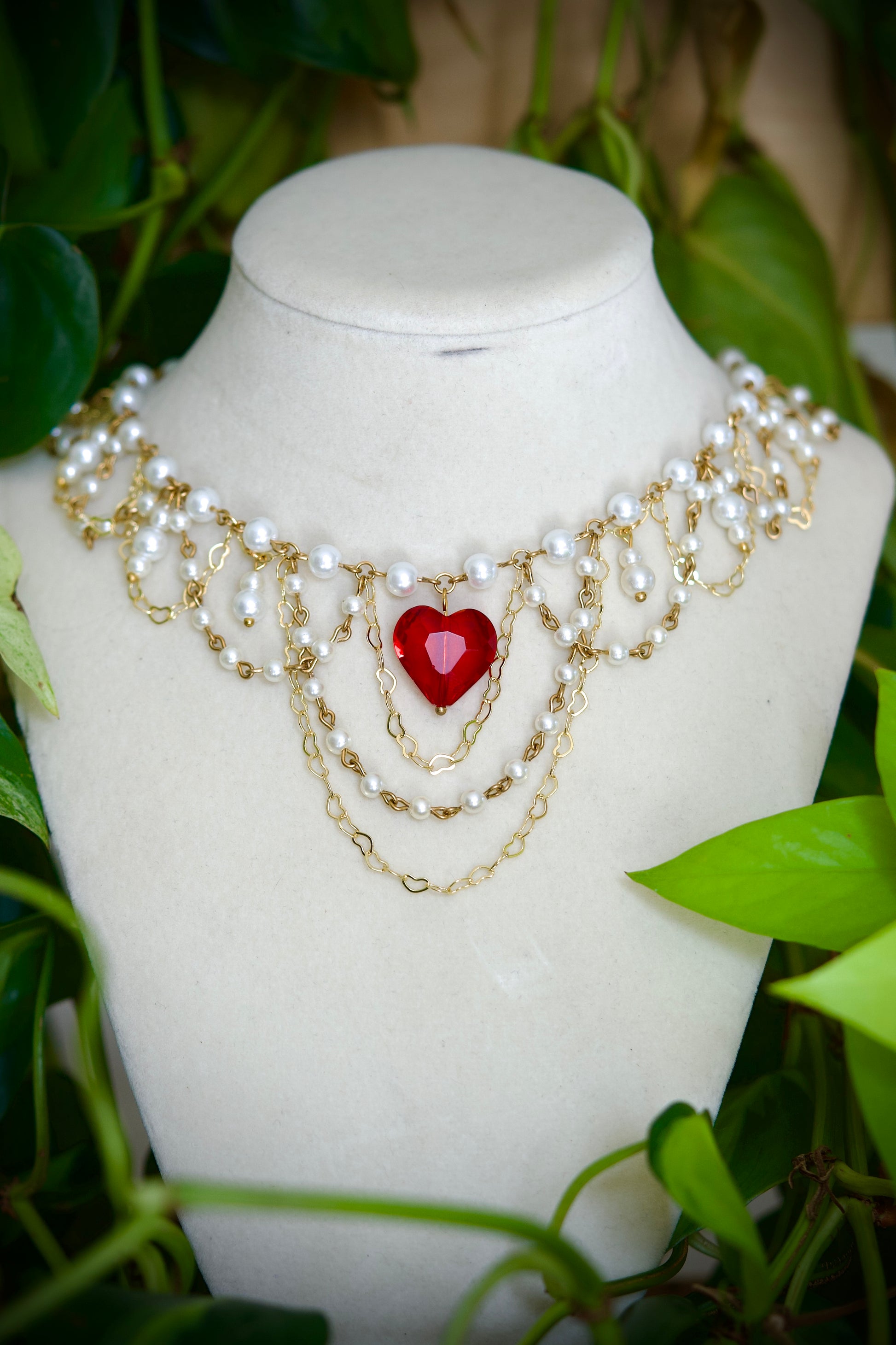 A gold and white pearl choker necklace with a central red heart charm, displayed on a white mannequin bust surrounded by green foliage.